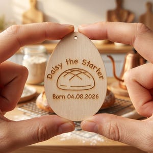 May include: A wooden, egg-shaped tag with the text "Daisy the Starter" and "Born 04.08.2026" engraved on it. The tag features a bread loaf illustration. The tag has a small hole at the top for hanging.