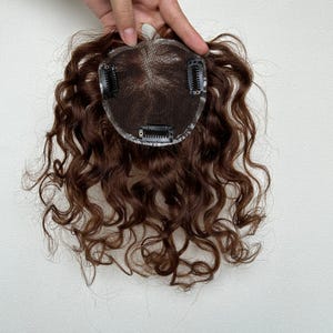 以下が含まれることがあります： 円形のメッシュベースに3つの黒いクリップが付いたブラウンのヘアトッパー。髪はルーズで明確なカールにスタイリングされており、中央のベースから外側に向かって層状に広がっています。