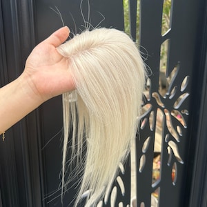 Puede incluir: Una pieza de cabello rubio claro con una textura fina y de aspecto natural. La pieza de cabello se sostiene, mostrando el color y el estilo. La pieza de cabello parece estar hecha de cabello real, diseñada para mezclarse con el cabello natural.