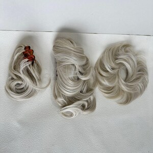 May include: Three hair extensions in a light platinum blonde shade. One piece features a tortoiseshell claw clip, another is a long wavy extension, and the third is a circular bun-style hairpiece with voluminous, soft, layered curls.