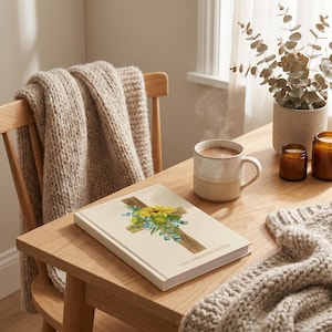 May include: A cream journal with the text "Lily's Prayer Notes" features a watercolour cross design with yellow sunflowers and blue accents. A mug of hot drink sits nearby on a wooden table, alongside a cosy blanket and candles.