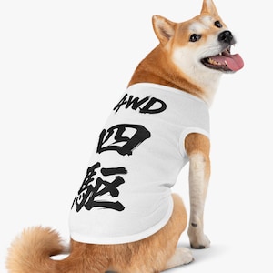 May include: A Shiba Inu dog wearing a white dog shirt with black Japanese characters and the letters "AWD" printed on it. The dog has reddish-brown fur and is sitting with its tongue out.