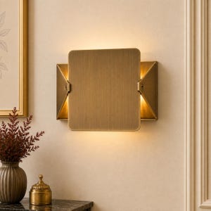 May include: A wall-mounted light fixture featuring a brushed brass finish with a square central plate and triangular side panels. The design emits a warm, soft glow from behind the central plate, illuminating the wall surface with light.