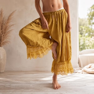 May include: Mustard-colored wide-leg pants with a drawstring waist and lace trim at the ankles. The pants are made of a lightweight fabric and have a flowing silhouette. The model is standing on one foot.