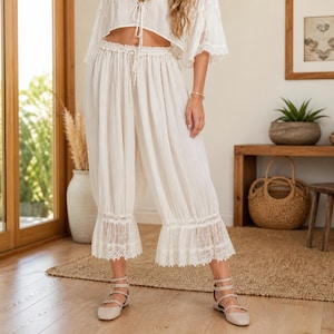 May include: Off-white, cropped top and matching wide-leg pants. The top has a tie-up front and short sleeves. The pants feature a drawstring waist and lace trim at the hem. The outfit is paired with beige flats. The setting is a well-lit room with wooden floors.
