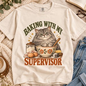 Könnte beinhalten: Ein cremefarbenes T-Shirt mit einer Grafik einer flauschigen grauen Katze in einer Rührschüssel. Der Text lautet BAKING WITH MY SUPERVISOR. Um die Katze herum sind ein Glasbehälter, zwei Eier, ein Stück Butter und ein Messbecher abgebildet.