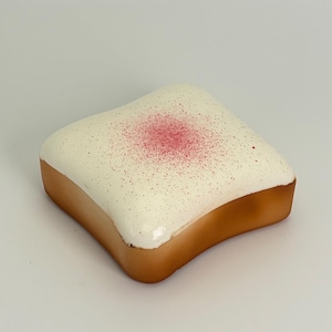 May include: A square-shaped pastry with a golden-brown crust and a smooth white icing top. The center of the white icing features a concentrated dusting of fine pink powder, which gradually fades into smaller, scattered pink specks across the surface.