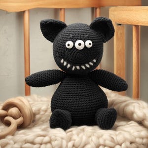 May include: A black crocheted monster toy with three white eyes and a stitched smile with white teeth. The handmade plushie has small arms and legs and is sitting on a cream-colored blanket. The toy is a unique, handmade item.