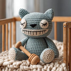 May include: A handmade, crocheted blue cat-like plush toy with a wide, stitched grin and closed eyes. It holds a wooden honey dipper. The toy is sitting on a cream-colored, textured blanket.