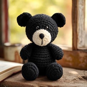 May include: A handmade black crochet teddy bear with a cream-colored muzzle and a black nose. The bear has small black eyes and large, rounded ears. It is sitting on a wooden surface near an open book and a window.