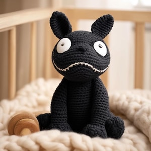 May include: A handmade black crochet creature with large white eyes and a stitched grin. The plush toy has long ears and is seated on a soft, cream-colored blanket. A wooden toy is partially visible in the foreground, suggesting a nursery setting.