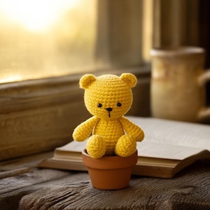 May include: A handmade yellow crochet teddy bear sits in a small terracotta pot. The bear has black eyes and a black nose. The background includes a window, a book, and a mug, all resting on a wooden surface.