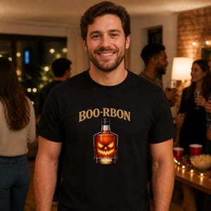 Peut inclure: Un t-shirt noir à manches courtes avec un motif représentant une bouteille en verre remplie d'un liquide ambré. La bouteille présente un visage de citrouille d'Halloween orange lumineux. Le texte BOO-RBON est imprimé en lettres dorées.