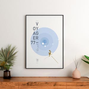 May include: A framed wall art print featuring a minimalist graphic design of the Voyager 77 space probe. The poster displays concentric light blue circles, a gold-toned illustration of the spacecraft, and the vertical text VOYAGER 77.