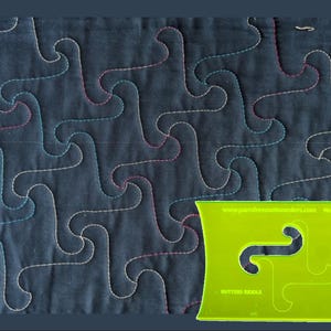 May include: A lime green acrylic quilting template labeled "BUTTERS RIDDLE" and "No 15b" with a central cutout shape, positioned over a dark fabric background featuring a repeating, interlocking stitched pattern in light blue and pink thread.