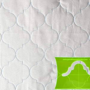 May include: White quilted fabric with a repeating quatrefoil pattern, stitched with light blue thread. A green acrylic quilting ruler with a matching design is in the lower right corner. The ruler has the text "www.parrsfreemotionrulers.com No.9b".