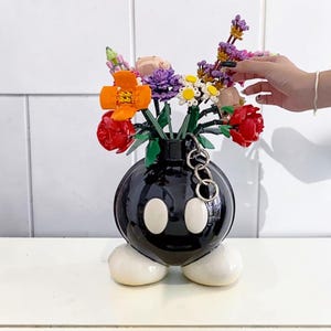 May include: A black ceramic vase in the shape of a cartoon bomb, featuring white oval eyes and feet. The vase is filled with a vibrant assortment of artificial flowers, including red, orange, purple, and white. A silver chain is attached.