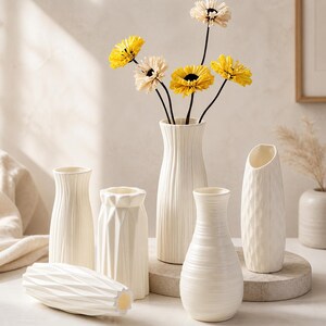 May include: A collection of white ceramic vases in various shapes and sizes, some with textured surfaces. Yellow and cream-colored artificial flowers are arranged in one vase. The vases are displayed on a neutral surface.