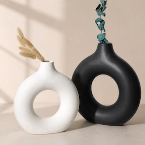 May include: Two modern ceramic vases, one white and one black, each with a circular design. The white vase holds dried beige grass, while the black vase contains teal-colored decorative stems. The vases have a minimalist aesthetic.