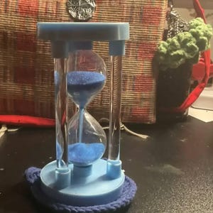 May include: A blue hourglass with clear glass chambers filled with blue sand. The hourglass is set on a blue base with a blue knitted coaster. The hourglass is a decorative item.
