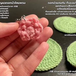 Puede incluir: Un llavero de flor de ganchillo rosa hecho a mano y tres posavasos de hoja de nenúfar de ganchillo verde. La flor se sostiene en una mano. Las hojas de nenúfar miden aproximadamente 8 cm de diámetro y 0,3 cm de grosor. El texto está en tailandés.