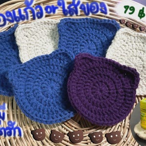 May include: A selection of handmade crochet coasters in blue, white, and purple, displayed on a woven basket. The coasters are square with rounded corners. A small jar of yellow ointment and keys are also visible.