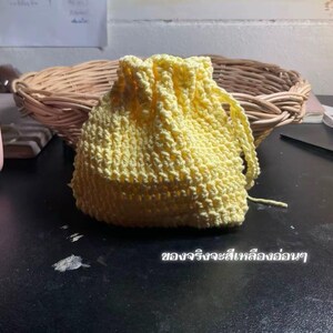 以下が含まれることがあります： 柔らかい黄色で作られた、手作りの小さな巾着袋。バッグはテクスチャードステッチでかぎ針編みされており、上部は巾着でまとめられています。背景には柳のバスケットと暗い面が含まれています。