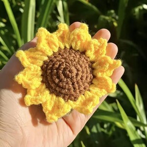 Puede incluir: Un girasol de ganchillo hecho a mano con pétalos amarillos brillantes y un centro marrón. La flor se sostiene en una mano, con follaje verde en el fondo. Este artículo artesanal es un acento floral decorativo.