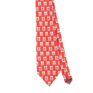 May include: A bright red necktie featuring a repeating pattern of white, square-shaped motifs. Each white square contains a small, centred blue and black graphic design. The tie has a pointed tip and a smooth, uniform texture throughout the fabric.