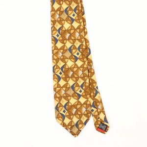 Peut inclure: Une cravate jaune avec un motif géométrique répétitif composé de carrés bruns arrondis, d'accents courbes bleus et de petits losanges blancs. L'étiquette noire en bas porte le texte blanc 100% SILK MADE IN U.S.A. FABRIC IMPORTED FROM ITALY.
