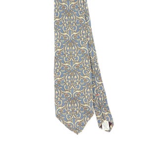 May include: A blue necktie featuring a repeating intricate gold and cream-colored scrollwork pattern. The tie has a pointed end and a white fabric care label attached to the back, which includes printed text regarding material and cleaning instructions.
