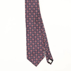 May include: A necktie featuring a dark blue background with a repeating geometric pattern of red and white diamond shapes. Each diamond contains a small, intricate blue and white paisley-style motif. The tie has a pointed end and a label.