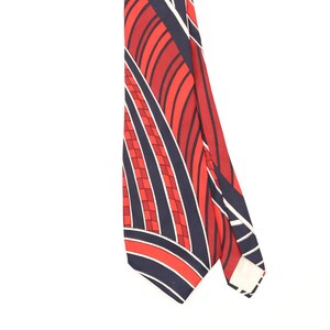 Puede incluir: Una corbata con un patrón geométrico audaz de rayas diagonales alternas en azul marino profundo y rojo vibrante. El diseño incluye líneas blancas finas y una sección con un patrón de cuadrícula de pequeños cuadrados en tonos rojos.