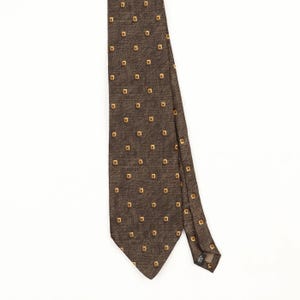 May include: A brown necktie featuring a repeating pattern of small, light gold-coloured squares. The fabric has a visible textured weave, and the tie is shown hanging vertically against a plain white background, displaying its pointed end.