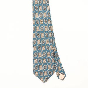 Pode incluir: Uma gravata azul com um padrão repetitivo de formas retangulares e ovais na cor creme com detalhes em linhas castanho-escuras. A gravata tem uma extremidade pontiaguda e uma etiqueta preta visível na parte de trás, com design geométrico.