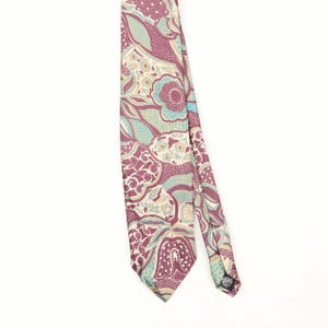 Puede incluir: Una corbata con un patrón complejo y multicolor de flores estilizadas, formas de cachemira y texturas geométricas en tonos rosa polvoriento, verde azulado claro, crema y granate oscuro. La tela tiene un acabado liso y borde inferior.