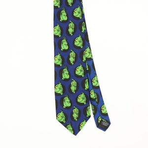 Op de afbeelding: Een blauwe das met een herhalend patroon van groene stripfiguurgezichten in zwarte cirkels. De das heeft een puntig uiteinde en een zwart stoffen label aan de achterkant bij de onderkant, met witte tekst.