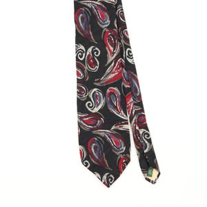 May include: A black necktie featuring a repeating paisley pattern in shades of red, blue, and grey. The fabric has a smooth finish, and a small green tag near the pointed tip displays the text ALL SILK in white capital letters.