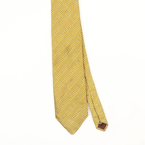May include: A mustard yellow necktie featuring a repeating pattern of diagonal wavy lines in white and dark grey. The tie has a pointed end and a visible dark label on the underside of the narrow tail, showcasing a consistent geometric design.