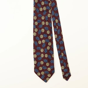 May include: A burgundy necktie featuring a repeating pattern of circular tan shapes and blue fan-like motifs. The tie has a pointed end and a black label on the back that reads 100% SILK MADE IN ITALY, displaying a classic design and construction.