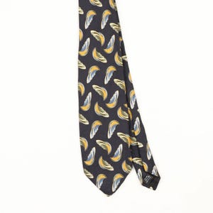 May include: A dark navy blue necktie featuring a repeating pattern of stylised crescent shapes in shades of golden yellow, light blue, and white. The tie has a pointed tip and a black fabric label attached to the back side near the bottom edge.