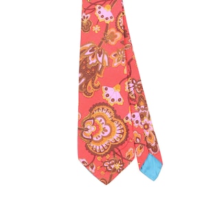 Pode incluir: Uma gravata com um fundo vermelho-coral vibrante e um padrão repetitivo de flores e paisley em tons de castanho, laranja, amarelo e rosa claro. A gravata tem uma extremidade pontiaguda e um forro azul brilhante visível na borda dobrada.