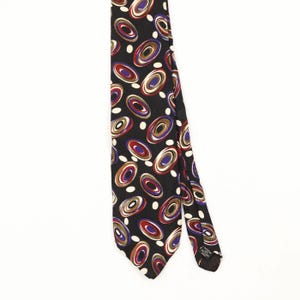 Puede incluir: Una corbata negra con un patrón repetitivo de formas ovaladas concéntricas en tonos rojo, azul, blanco y marrón. El diseño incluye pequeños puntos blancos intercalados entre los óvalos. Una pequeña etiqueta de cuidado negra está unida al reverso.