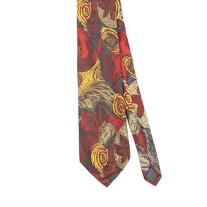Puede incluir: Una corbata con un patrón abstracto de formas circulares arremolinadas y figuras orgánicas en tonos rojo intenso, amarillo brillante y beige apagado sobre un fondo gris carbón oscuro, con una punta y un extremo posterior doblado.