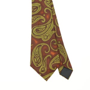 May include: A necktie featuring a dark brown background with a repeating light olive green paisley pattern. The fabric has a smooth texture and a pointed tip, with a dark grey lining visible on the underside of the folded section of the tie.