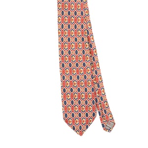 Peut inclure: Une cravate présentant un motif géométrique répétitif dans des tons de rouge, bleu marine et or. Le design se compose de formes ovales et festonnées interconnectées. Une petite étiquette rectangulaire avec le texte "TRENT" est visible au dos.