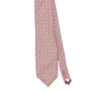 May include: A necktie featuring a repeating paisley pattern in shades of red, light blue, and white. The fabric displays a dense, intricate design of teardrop-shaped motifs across the entire surface, with a pointed end and a visible fabric label.