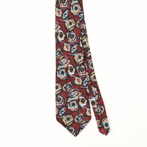 May include: A necktie featuring a deep red background with an abstract pattern of geometric shapes in shades of tan, light blue, and dark brown. The design consists of repeating, irregular square and circular motifs distributed across the fabric.