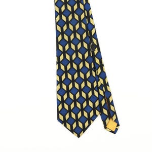 Puede incluir: Una corbata con un patrón geométrico repetitivo de rombos azules y amarillos delineados en negro. La tela muestra un efecto de cubo tridimensional estructurado en toda la superficie, con un forro amarillo sólido visible en la punta inferior.