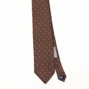 Peut inclure: Une cravate marron ornée d'un motif répétitif de petits carrés blancs. Le tissu présente un tissage texturé et comporte une petite étiquette grise au dos indiquant All Silk. La cravate est présentée verticalement sur un fond blanc.